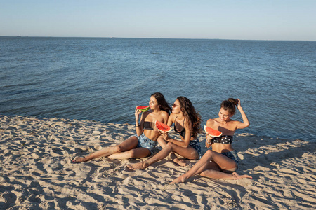 三个穿着牛仔裤短裤和黑色胸罩的漂亮女孩坐在海边的沙滩上, 拿着西瓜片