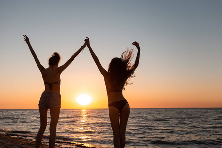 在日落时, 两个穿着泳衣的身材苗条的黑发女孩举起手来