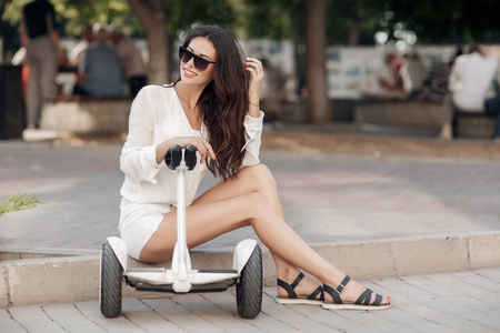 穿着白色衬衫和短裤的漂亮女人在夏天在城市公园户外骑着一辆Segway。年轻的黑发女孩在绿色公园里带着电动迷你悬停板滑板车。 好天