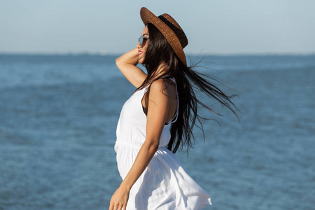 在一个阳光明媚的日子里, 在海边, 穿着白色礼服太阳镜和棕色帽子的黑发女孩的肖像