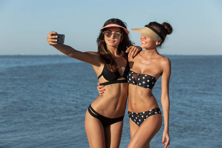 在一个阳光明媚的日子里, 两个穿着泳衣头戴帽子的热辣的黑头发女孩微笑着, 在海边自拍自拍