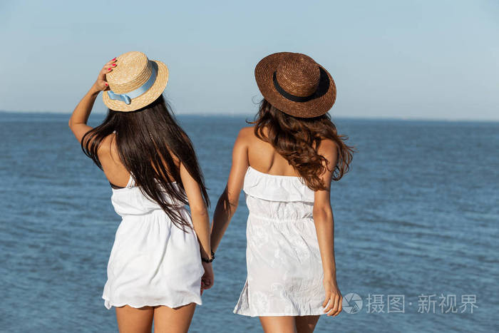 在一个阳光明媚的日子里两个穿着白色衣服头戴帽子的黑发女孩站在海边