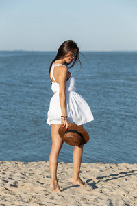 在阳光明媚的日子里, 穿着白色礼服戴着太阳镜的迷人黑发女孩手里拿着帽子, 走在海边的沙滩上