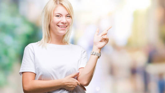 年轻漂亮的金发女人穿着白色T恤，在孤立的背景上，脸上挂着一个大大的微笑，用手和手指指向一边，看着相机。