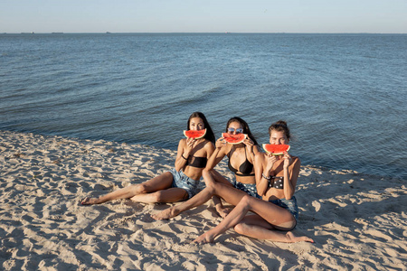 三个穿着牛仔裤短裤和黑色胸罩的漂亮女孩坐在海边的沙滩上, 把西瓜片放在嘴唇附近