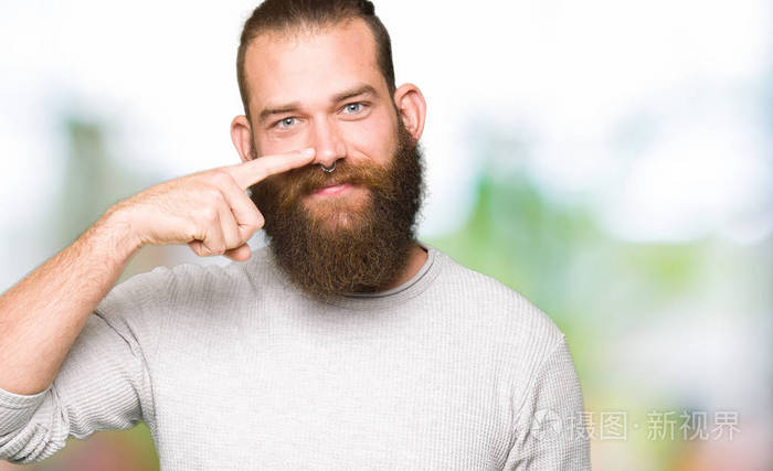 年轻的金发男子穿着休闲毛衣用手指着脸和鼻子笑容开朗