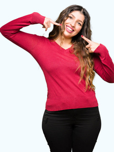 年轻漂亮的女人穿着红色毛衣，微笑自信，用手指牙齿和嘴指着。健康观念。