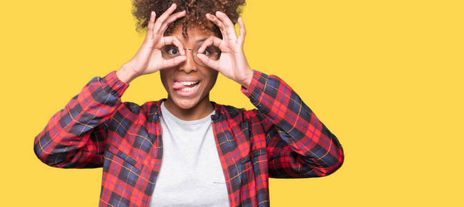 美丽的年轻非洲裔美国妇女戴着眼镜，在孤立的背景下做着好的手势，就像望远镜伸出舌头，透过手指看眼睛。 疯狂的表情。