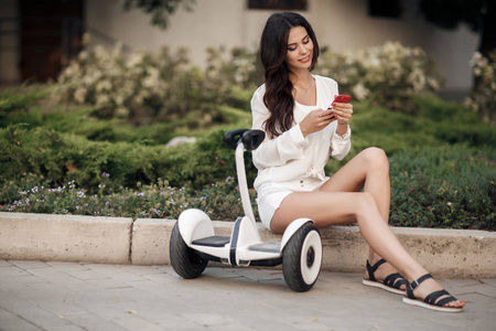 穿着白色衬衫和短裤的漂亮女人在夏天在城市公园户外骑着一辆Segway。年轻的黑发女孩在绿色公园里带着电动迷你悬停板滑板车。 好天