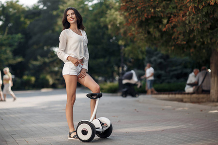 穿着白色衬衫和短裤的漂亮女人在夏天在城市公园户外骑着一辆Segway。年轻的黑发女孩在绿色公园里带着电动迷你悬停板滑板车。 好天