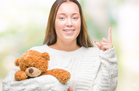 年轻的白种人女人抱着泰迪熊，在孤立的背景下惊讶地发现了一个想法或问题，手指着幸福的脸第一