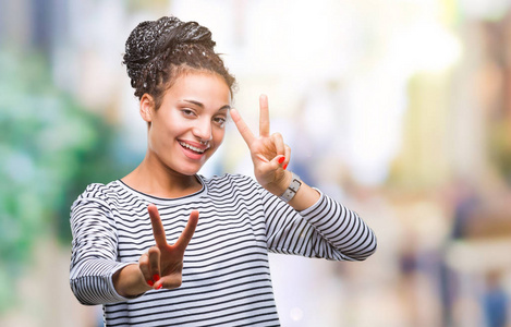 年轻的辫子非洲裔美国女孩穿着毛衣，在孤立的背景上微笑，看着相机，露出手指，做胜利标志。 二号。