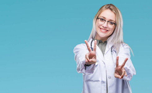 年轻的金发医生女人在孤立的背景下微笑着看着镜头，显示手指在做胜利标志。 二号。