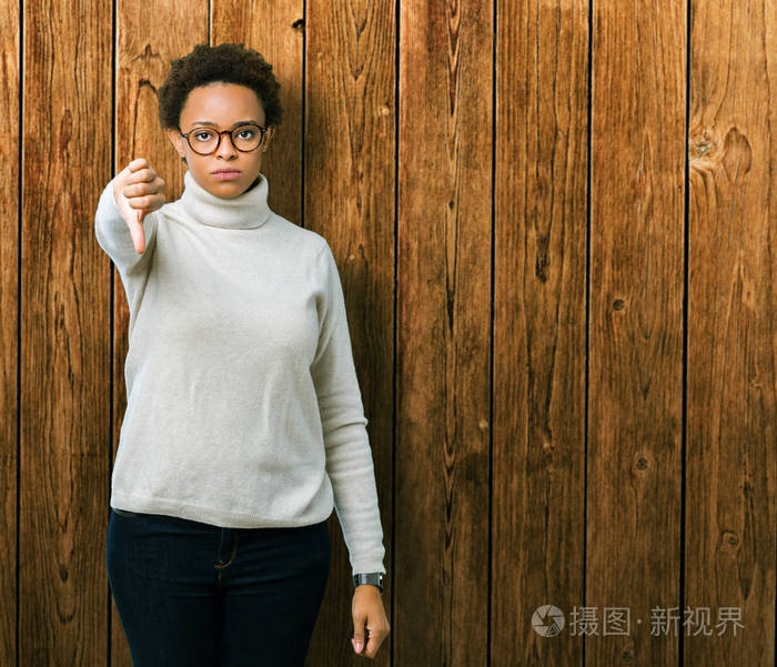 年轻美丽的非裔美国妇女戴着眼镜，在孤立的背景上看起来不高兴和愤怒，表现出拒绝和消极的拇指向下的手势。 不好的表情。