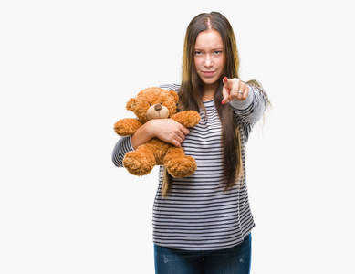 年轻的白种人女人拿着泰迪熊在孤立的背景上指着相机和你的手，从前面签署积极和自信的手势。