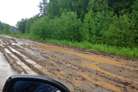 从汽车上观看可怕的乡村土路，夏天的时候水坑和泥被绿色的森林包围。 俄罗斯肮脏破碎的乡村道路