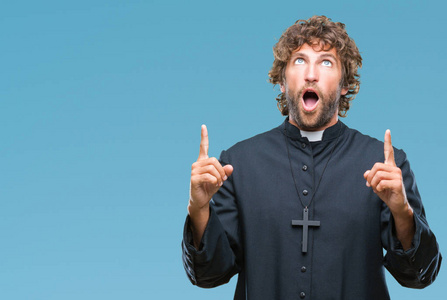 英俊的西班牙裔天主教牧师在孤立的背景下惊讶和惊讶地抬头，用手指和举起的手臂指着。