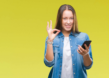 年轻美丽的高加索女人用智能手机在孤立的背景下发送信息，用手指做OK标志，非常好的符号