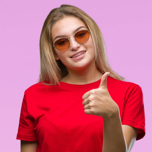年轻的高加索女人戴着太阳镜，在孤立的背景下，用手做快乐的竖起大拇指的手势。 赞许的表情看着相机，显示出成功。