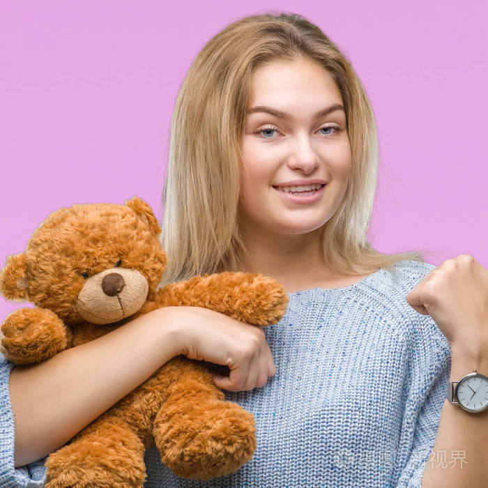 年轻的白种人女人抱着可爱的泰迪熊在孤立的背景上指着用拇指向旁边
