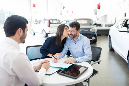 高兴的女人亲吻男人在汽车经销商签订买卖协议