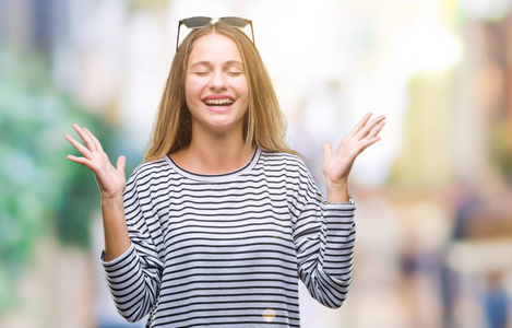 年轻漂亮的金发女人戴着太阳镜在孤立的背景上庆祝疯狂和疯狂的成功，举起手臂和闭上眼睛尖叫兴奋。 赢家概念