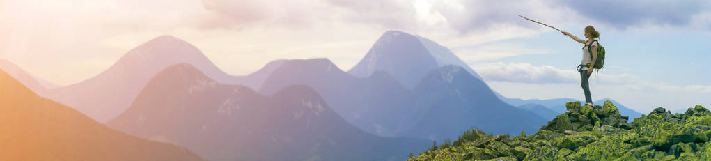 年轻的苗条金发旅游女孩与背包点与棍子在雾山山脉全景站在岩石顶部明亮的蓝色早晨的天空背景。 旅游旅游和攀岩概念。