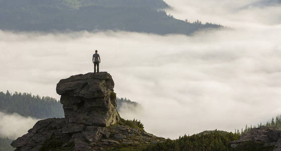 运动登山游客在高山山谷高岩石组的轮廓，充满白色的蓬松云和雾，并覆盖着常绿森林山坡在晴朗的天空背景下。