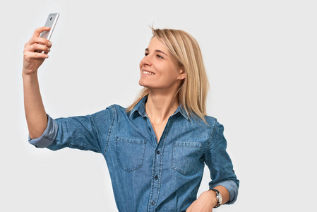 快乐的金发女性高兴地看着智能手机的相机，自拍穿着牛仔衬衫，在白色的工作室背景下摆姿势。 积极的学生妇女在手机上拍摄自我肖像