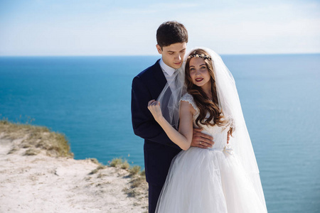 新婚夫妇的肖像在悬崖，美丽的海景。 穿着优雅西装的新郎拥抱穿着白色裙子的新娘。 刚结婚的人
