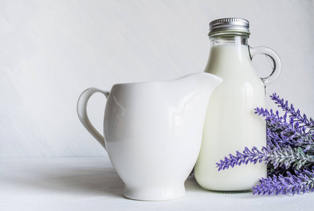带有牛奶的老式瓶子，旁边是一个漂亮的水壶，白色复古背景上有一枝薰衣草花