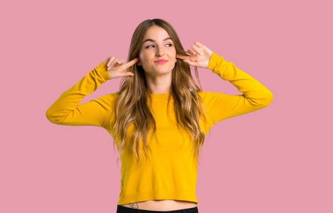 一个年轻的女孩，穿着黄色毛衣，双手遮住两只耳朵，孤立的粉红色背景