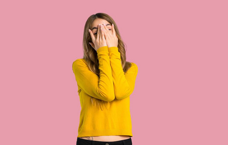 年轻的女孩，穿着黄色毛衣，双手遮住眼睛，用手指看着孤立的粉红色背景