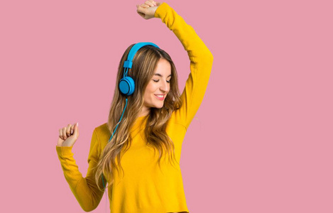 带着黄色毛衣的年轻女孩带着耳机听音乐，在孤立的粉红色背景上跳舞