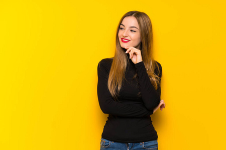 黄色背景的年轻漂亮女人看着一边,手放在下巴上照片