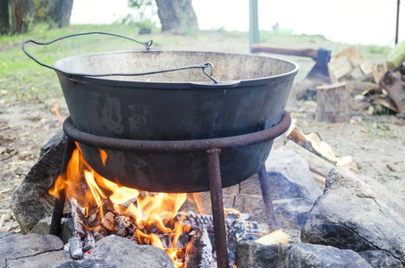 大金属锅，盖子和手柄在三脚架上，用于烹饪。 在大自然的火堆上用礼帽做饭