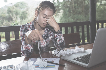 工作失败概念商务女性头痛，而工作使用笔记本电脑。 压力和沮丧的女孩摸着她的头，感到疼痛，而坐在木桌旁。