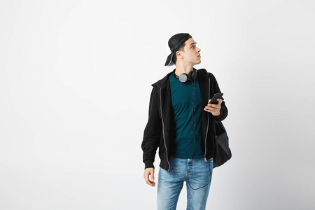 一个背着黑色背包的家伙, 脖子上戴着耳机, 穿着深色 t恤牛仔裤运动衫和帽子, 在演播室里用手机在白色背景下使用手机