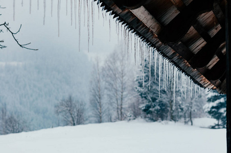 长长的明亮的透明的冰柱，贴着薄雾般的森林，挂在木屋顶上。 选择性聚焦。 背景中的雪山和树木
