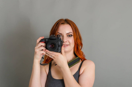 带着红色头发的微笑的女人手里拿着现代相机。 灰色背景创意摄影师