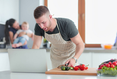 年轻男子切蔬菜和女人站在厨房里的笔记本电脑