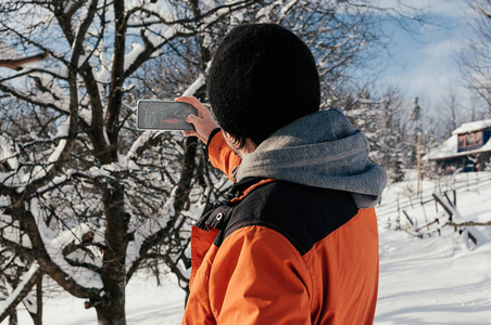 年轻人用智能手机橙色服装拍照的背面视图。 冬日雪树乡村。 新鲜空气。 乐趣及休闲概念