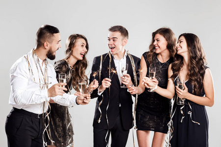 公司的女孩和男人穿着时尚优雅的衣服有乐趣在一起, 拿着香槟的眼镜和闪闪发光的手在白色背景的糖果周围。派对时间