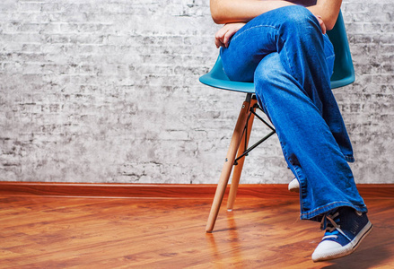 穿着牛仔裤和运动鞋的女性腿坐在灰色墙壁背景的椅子上,有复印空间