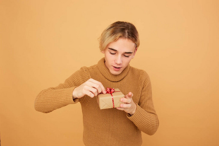 穿着芥末色毛衣的年轻金发碧眼的家伙拿着一个小礼物, 手里拿着红色的弓在工作室的米色背景
