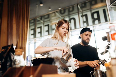 在一家舒适的咖啡店里, 两个年轻的咖啡师, 一个金发碧眼的女孩, 一个留胡子的时尚男人, 一起在咖啡机里煮咖啡