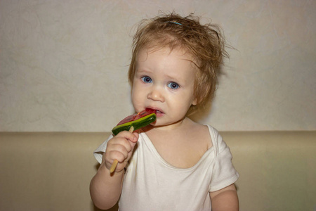 一个衣衫不整的蓝眼睛小女孩，穿着白色的身体，在棍子上尝糖果，棒棒糖，一个三角形的西瓜。