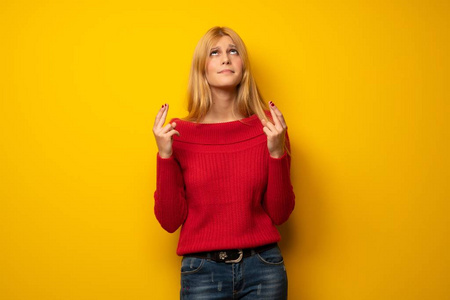 金发女人越过黄色的墙，手指交叉，祝你一切顺利