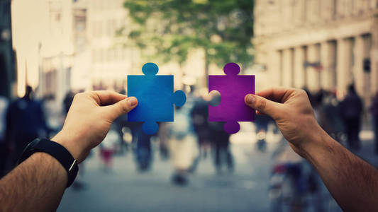两只手拿着不同的彩色拼图，在拥挤的街道背景上联合拼图。 业务解决方案的成功和策略多样性连接概念。 团队合作小组合作。