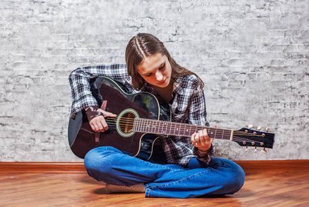 年轻的少年黑发女孩，长发坐在地板上，在灰色的墙壁背景上弹着黑色的吉他